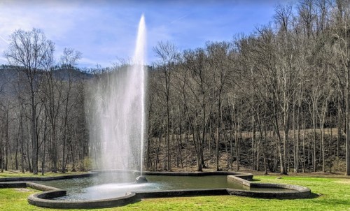 A tall fountain sprays water into the air, surrounded by a grassy area and bare trees against a blue sky.