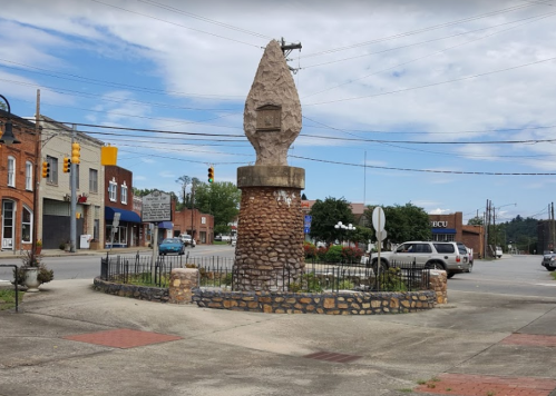 A stone monument shaped like a flame, surrounded by a circular stone base, located in a small town intersection.
