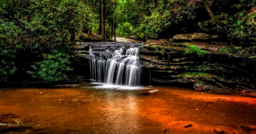 A serene waterfall cascades over rocky ledges into a tranquil, reddish-brown pool, surrounded by lush greenery.