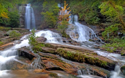 A serene landscape featuring multiple waterfalls cascading over rocky terrain, surrounded by lush greenery and autumn foliage.