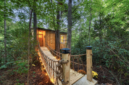 A cozy wooden cabin nestled in a forest, connected by a rope bridge illuminated with soft lights.