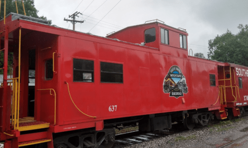 A bright red caboose with a logo, parked on a railway track under a cloudy sky.