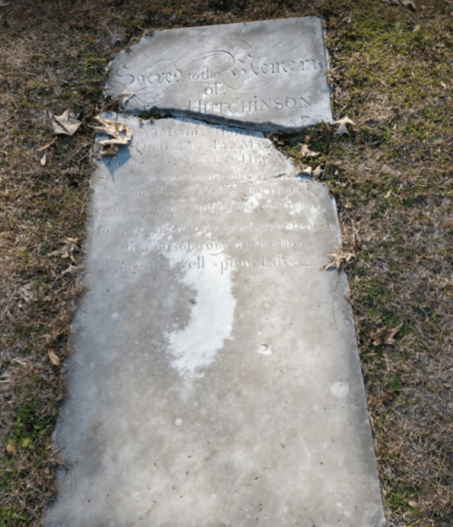 A weathered gravestone partially cracked, inscribed with "Sacred to the Memory of" and the name Hutchinson.