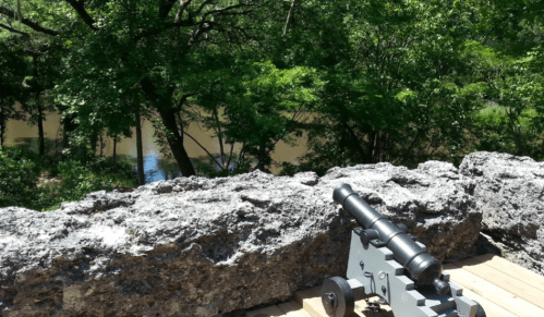 A cannon positioned on a rocky ledge overlooking a river surrounded by lush green trees.