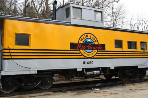 A yellow train caboose with "Main Line Rio Grande" logo and black stripes, parked on a railway track.