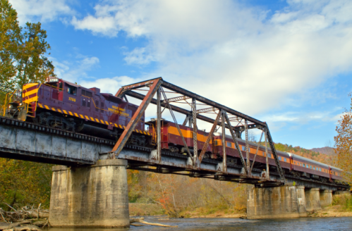 A vintage train crosses a metal bridge over a river, surrounded by trees and a blue sky with clouds.