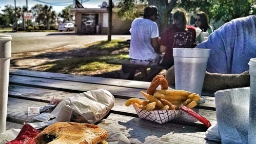 A table with food items, including fries and a sandwich, with people sitting in the background at a casual outdoor setting.