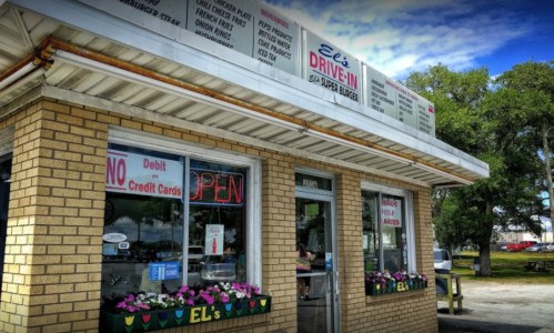Exterior of Eli's Drive-In featuring a brick facade, open sign, and menu board, with flowers in the window boxes.
