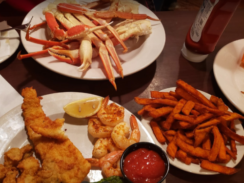 A table with two plates of seafood: crab legs, fried fish, shrimp, and sweet potato fries, with ketchup on the side.