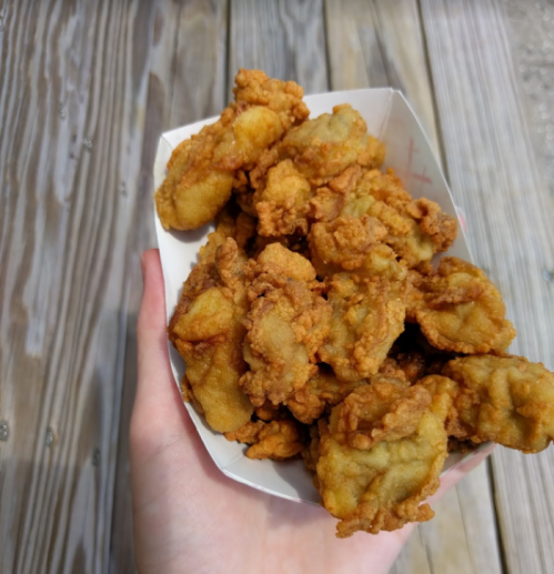 A hand holding a paper tray filled with golden-brown fried food, likely chicken or seafood, on a wooden surface.