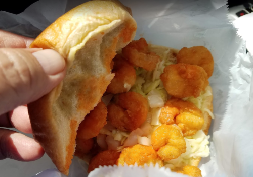 A hand holding a sandwich filled with crispy shrimp and lettuce, wrapped in white paper.