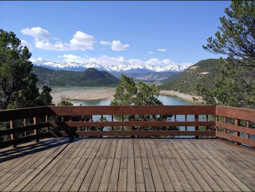 Scenic view from a wooden deck overlooking a lake and snow-capped mountains under a blue sky with fluffy clouds.