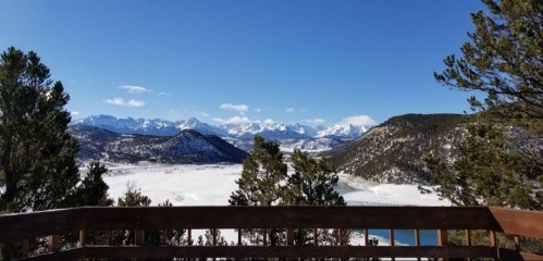 A scenic view of snow-covered mountains and a valley, framed by trees under a clear blue sky.
