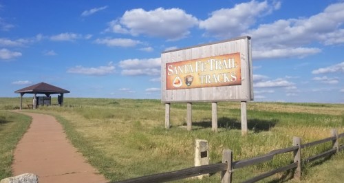 Sign for Santa Fe Trail Tracks with a path leading to a gazebo under a blue sky with fluffy clouds.