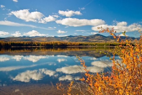 A serene lake reflects autumn foliage and blue skies, surrounded by mountains and fluffy clouds.