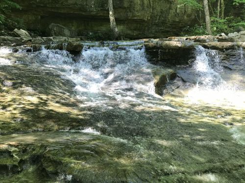 A serene waterfall cascades over rocks, surrounded by lush greenery and a rocky cliff in the background.