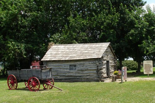 A rustic log cabin with a wooden wagon nearby, surrounded by green grass and trees. Informational sign in view.