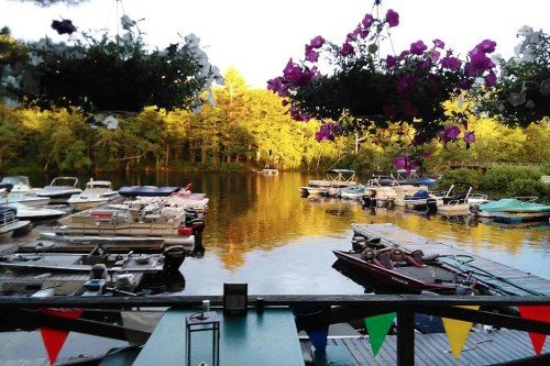 A serene lake scene with boats docked, surrounded by trees and hanging flower baskets, reflecting golden sunlight.