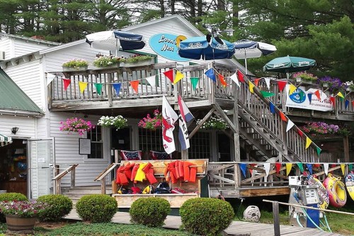A charming lakeside building with colorful flags, flower boxes, and outdoor seating, surrounded by greenery.