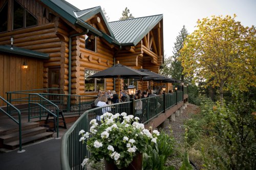 A log cabin restaurant with outdoor seating, surrounded by greenery and trees, during a warm evening.