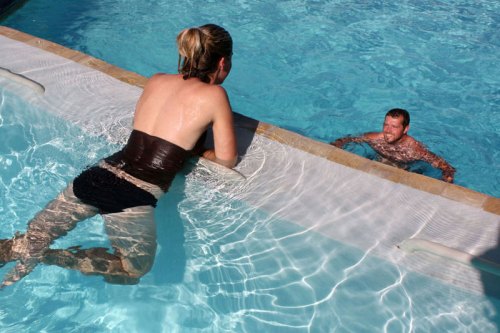 A woman in a swimsuit leans on the edge of a pool while a man swims nearby, both enjoying a sunny day.