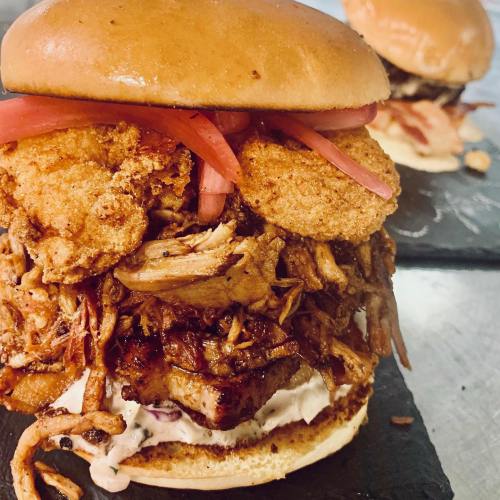 A close-up of a loaded burger with pulled pork, fried pickles, and pink onions on a soft bun, served on a black slate.