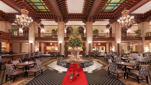 Elegant hotel lobby with a grand fountain, floral arrangements, chandeliers, and ducks on a red carpet.