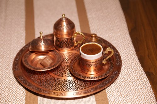 A decorative copper tray with a cup of coffee, sugar cubes, and ornate containers, set on a patterned tablecloth.