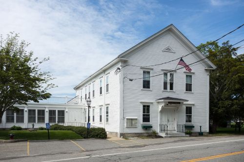 A white, two-story building with an American flag, surrounded by greenery and a parking area.