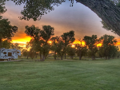 A serene campsite at sunset, featuring trees silhouetted against a colorful sky and an RV parked on the grass.