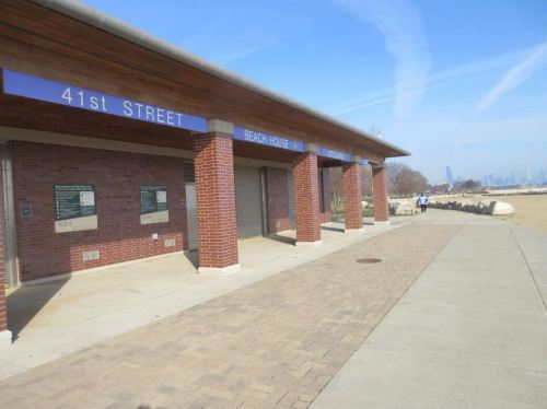 A brick building labeled "41st Street Beach House" next to a paved walkway, with a city skyline in the background.