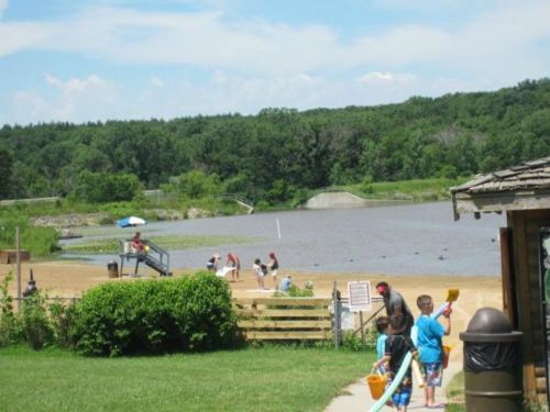 A sandy beach by a lake with children playing, surrounded by green trees and a clear blue sky.