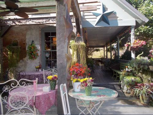 Charming outdoor café with colorful flowers, decorative plants, and cozy seating under a shaded porch.