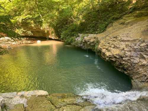 A serene natural scene featuring a clear blue pool surrounded by rocky cliffs and lush green trees.