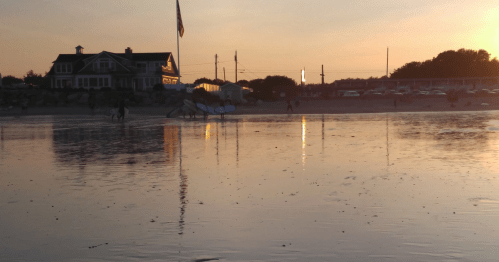 A beach at sunset with reflections on the water, a house in the background, and people walking near surfboards.