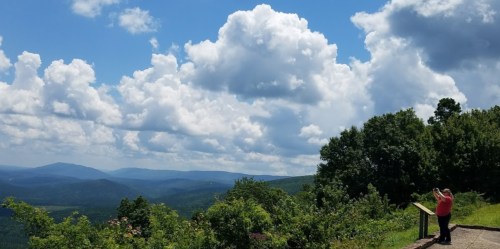 A person in a red shirt stands at a viewpoint, overlooking a lush green valley and a blue sky filled with clouds.