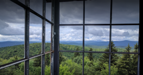 View from a tower window overlooking lush green mountains and a cloudy sky.