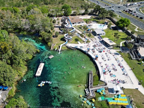 Aerial view of a water park with slides, a swimming area, and people enjoying the sun by a clear blue spring.