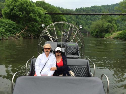 Two women sit on an airboat, wearing headsets, surrounded by lush greenery and a calm river.