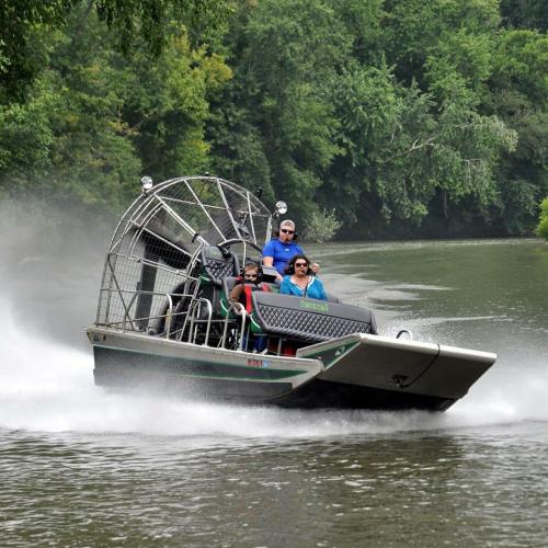 A group of people enjoying an airboat ride on a river, surrounded by lush greenery and splashing water.