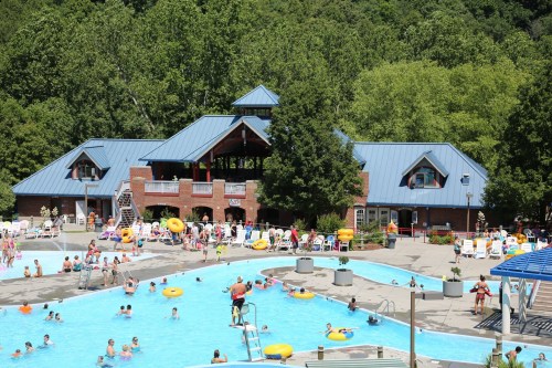 A busy outdoor pool area with people swimming, lounging, and enjoying the sun, surrounded by trees and a brick building.