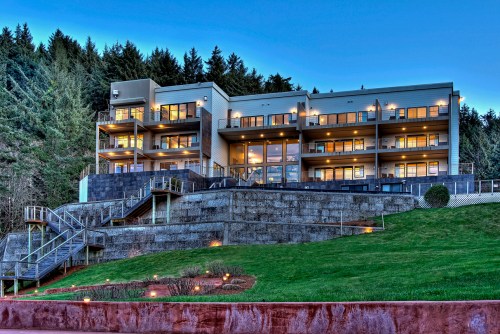 Modern multi-story building with large windows, surrounded by greenery and steps leading up to it, set against a forested backdrop.