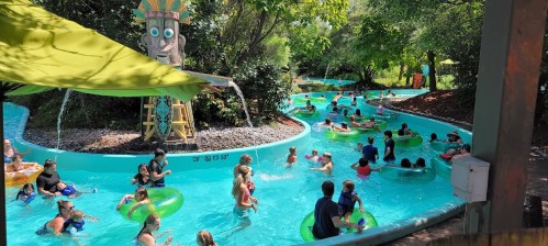 A busy lazy river at a water park, filled with people floating on tubes surrounded by greenery and playful water features.