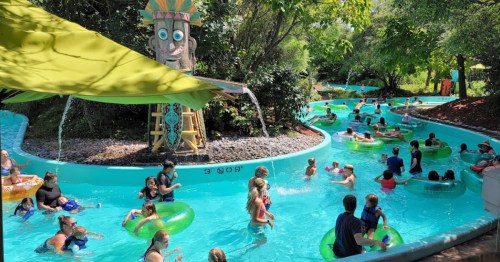 A busy water park lazy river with people floating on tubes, surrounded by greenery and a tiki statue.