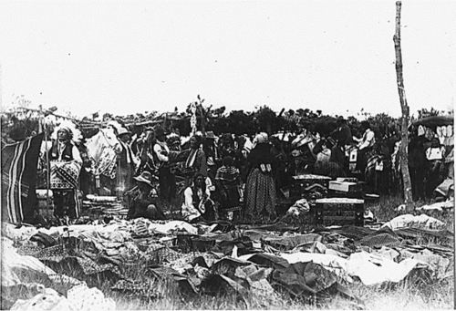 A historical scene depicting a gathering of people surrounded by various textiles and clothing in an outdoor setting.