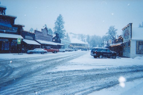 Snow-covered street in a small town, with shops and parked cars, creating a serene winter scene.