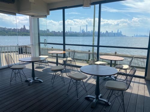 A bright indoor space with tables and chairs, overlooking a waterfront view of a city skyline and blue sky.