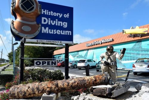 A diver statue waves in front of the History of Diving Museum, featuring a large diving helmet and a rusted anchor.