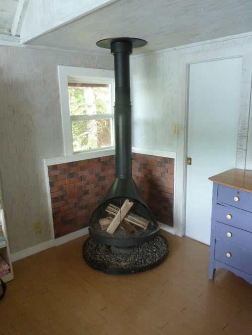 A modern black chimney fireplace with stacked wood, set in a cozy room with light-colored walls and a purple dresser.