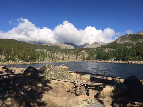 A serene lake surrounded by rocky shores, lush green trees, and mountains under a blue sky with fluffy clouds.